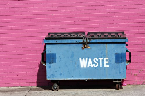 A large blue industrial rubbish bin with the word 'WASTE' in white capital letters is positioned on a concrete pavement against a bright pink painted brick wall. The bin has a metal lid with multiple ventilation slots and is equipped with black wheels at the base, allowing for movement. The surface of the bin shows signs of dirt and slight corrosion, indicating regular outdoor use. Shadows cast by the sunlight fall to the left side of the bin, suggesting a clear day. The surrounding environment appears to be an urban or commercial area, potentially part of a rubbish collection or waste management service in Brixton, with the vivid wall serving as a contrasting background to the bin, aligning with the context of commercial waste removal services offered by companies such as Commercial Waste Brixton.