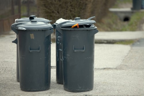 Two dark grey plastic rubbish bins with hinged lids are positioned on a paved driveway or street in an outdoor setting. The bins are cylindrical with smooth surfaces and small rectangular openings on their sides for ventilation or handling. One of the lids is slightly open, revealing an orange plastic object and some other miscellaneous waste inside. The background shows a blurred view of pavement, greenery, and a low bridge or culvert, indicating a suburban or urban environment possibly in Brixton. The surface of the bins shows minor scuffs and scratches, consistent with outdoor use. This image reflects typical waste collection containers used for rubbish disposal services in the local area, and it appears to be part of a collection or disposal process managed by Commercial Waste Brixton.