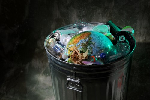 A close-up view of a metallic rubbish bin filled with various discarded plastic bottles, including water and soda bottles, some with transparent and greenish hues, and a crumpled piece of brown paper. The bin is positioned outdoors on a paved surface, with a dark, weathered wall in the background. The bottles are piled haphazardly, and a small globe map showing continents is visible among the waste, highlighting environmental concerns. The metallic surface of the bin shows subtle reflections and a latch secures the lid, which is open, revealing the mixed waste inside. This image relates to rubbish removal services offered by Commercial Waste Brixton, emphasizing waste collection and recycling efforts in the Brixton area, suggesting the importance of proper waste disposal in urban environments.