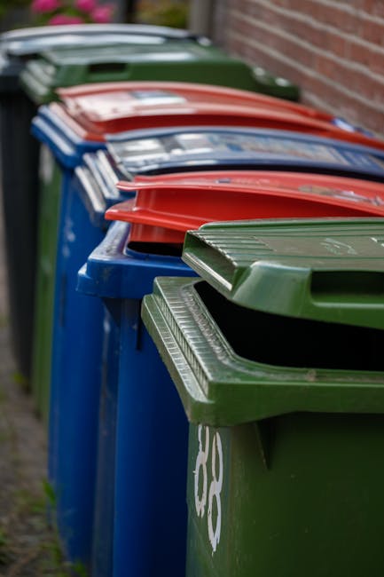 A tall blue plastic rubbish bin with a closed lid standing upright outdoors on a grassy area, with a yellow and blue sticker reading 'FIGHT BACK!' attached to its front. To the left, part of a red whe